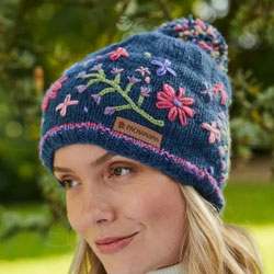 Pachamama Hampton Court Bobble Beanie. These Pachamama Bobble Beanies are knitted in Nepal by local villagers using local wools and natural dyes to UK designs. Each bobble beanie features a navy blue background with embroided pink, lilac and green flowers